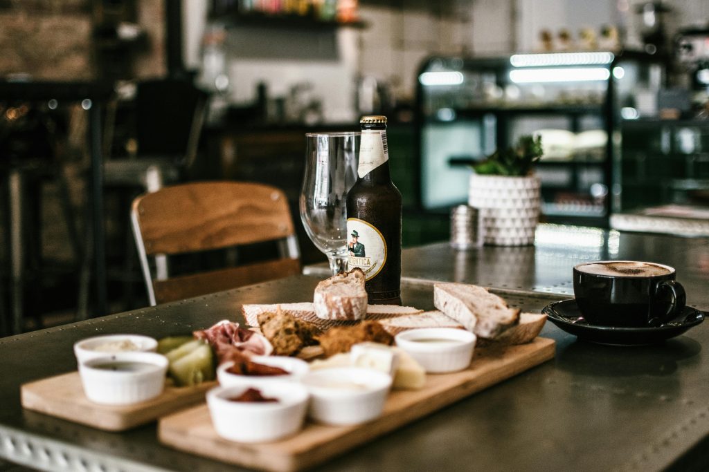 Rustic dining scene featuring craft beer, deli platter, and cappuccino in a cozy cafe atmosphere.