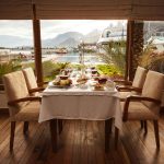 Elegantly set table in a seaside restaurant with a marina view, featuring gourmet dishes and fine dining setup.