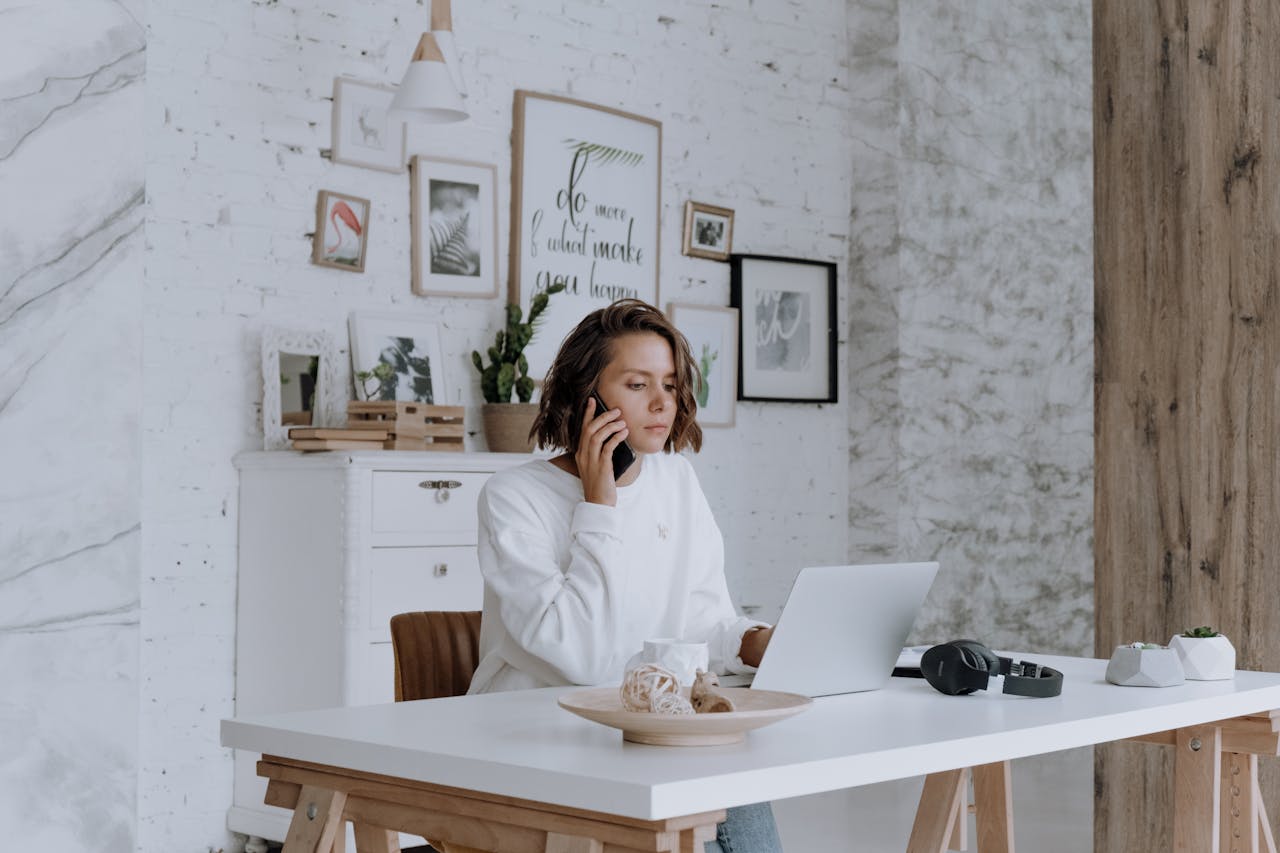 services-01 Woman talking on phone while using laptop in bright, modern home office with decor.