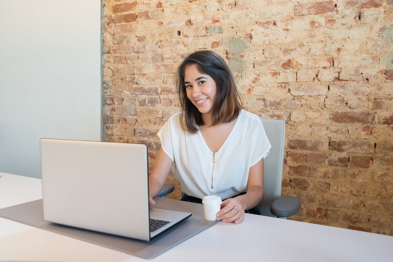 why-choose-us-02 A cheerful woman working on a laptop in a rustic office setting, holding a cup.