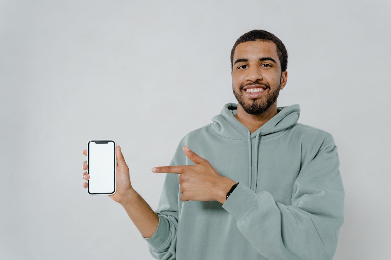 about-04 A smiling man in a hoodie points at a smartphone with a blank screen, ideal for mockup or design use.