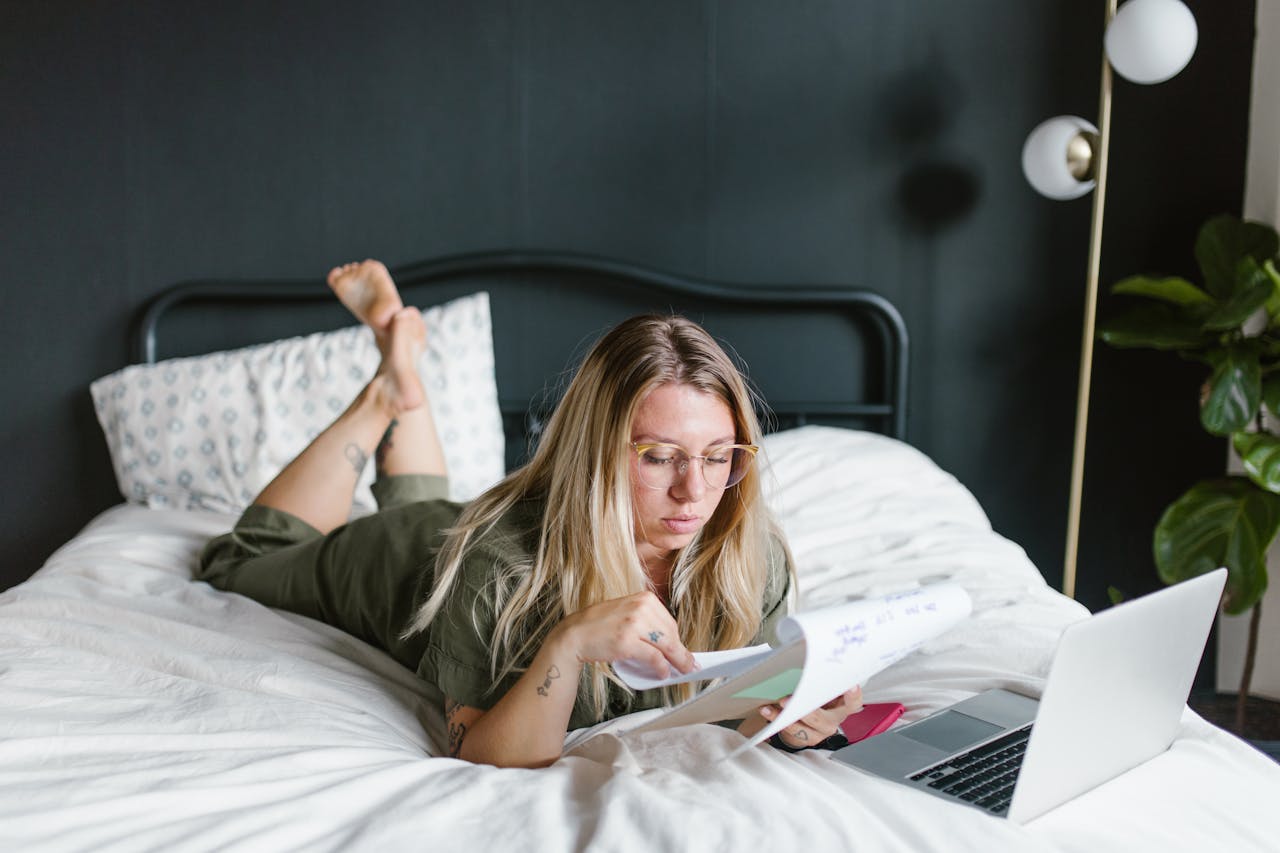 about-03 Young woman working from home, lying on bed with laptop and documents.