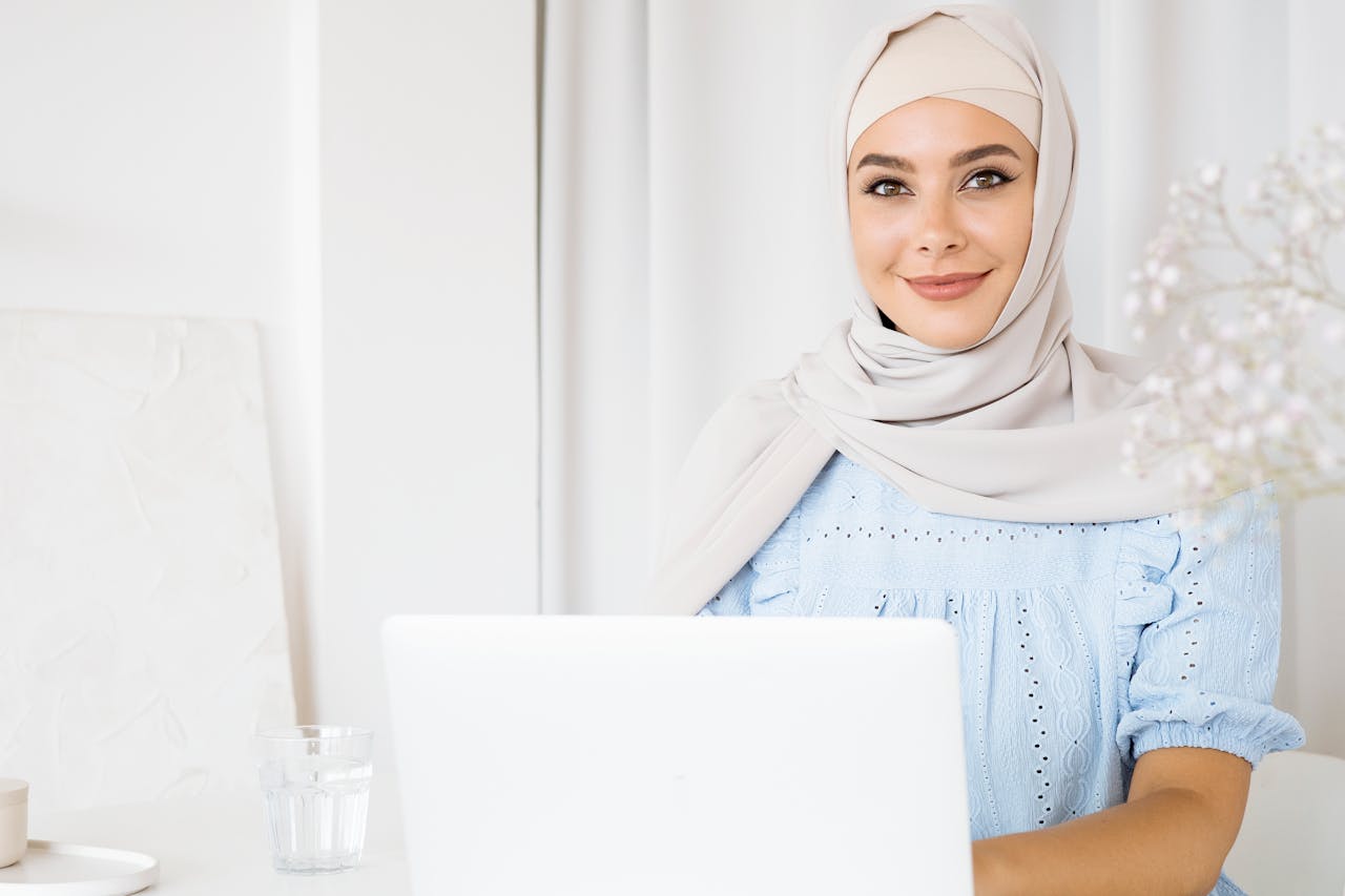 about-01 A smiling woman in a hijab using a laptop indoors with a bright and calm setting.